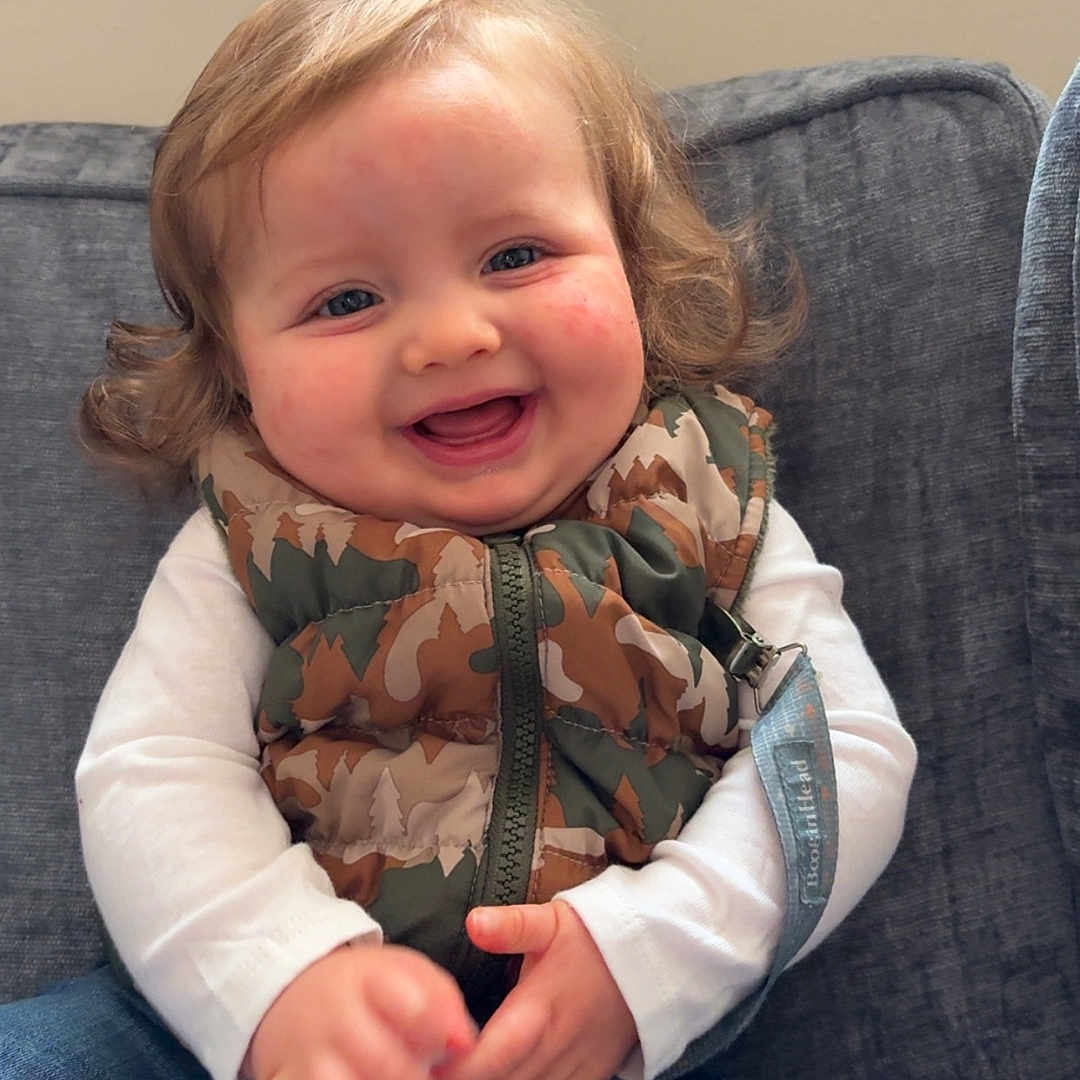 Kyson joined the competition — help win amazing prizes! toddler, child, smiling, happy, curly_hair, camouflage_vest, white_shirt, blue_jeans, sitting, couch, indoor, rosy_cheeks, face, person, clothing, casual, cute, young_child, portrait, cozy