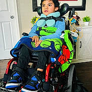 Aron is registered to the contest to win money with this photo: blanket, blue_clothing, carpet, child, curly_hair, door, expression, floor, furniture, home, indoor, neck_pillow, painting, person, plant, seated, sneakers, straps, support, wheelchair