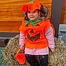 child, pumpkin_costume, orange, bucket, hay_bale, outdoor, seasonal, fall, halloween, smiling, black_shoes, leggings, pink_jacket, pigtails, festive, holiday, costume, cute, fun, celebration
