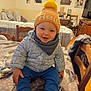 toddler, child, hat, pom_pom, jacket, scarf, smiling, table, chair, indoor, home, family_photos, wooden_furniture, cozy, patterned_tablecloth, happy, cute, warm_clothing, baby, portrait