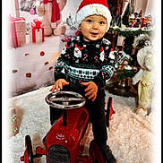 Linzo a rejoint le concours — aidez-le/la à gagner de superbes lots ! toddler, child, toy_car, christmas_hat, christmas_sweater, holiday_decorations, plush_bear, wrapped_gifts, white_rug, festive, indoor, smiling, seasonal, cute, red, black_pants, blue_eyes, decor, portrait, winter
