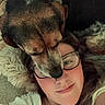 dog, person, face, glasses, lying_down, carpet, fur, close_up, affection, smile, nose_ring, indoor, casual_clothing, brown_fur, black_fur, white_clothing, cozy, resting, pet, human
