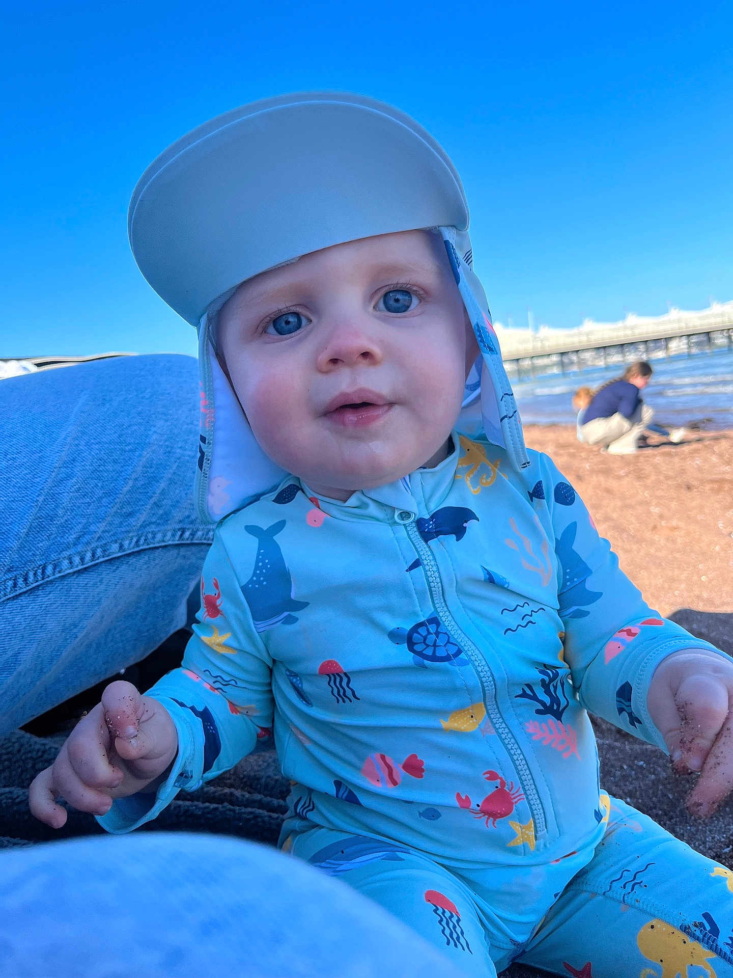Oaklyn-Jay joined the competition — help win amazing prizes! baby, child, beach, sand, blue_sky, swimsuit, hat, water, pier, playing, outdoor, sunny, cute, closeup, person, infant, sitting, marine_life_pattern, daytime, curious
