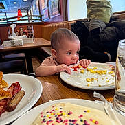 Anova is registered to the contest to win money with this photo: baby, bacon, breakfast, child, coat, curious, diner, food, glass_of_water, high_chair, indoor, ketchup, pancake, plate, sausage, scrambled_eggs, seat, sprinkles, table, window