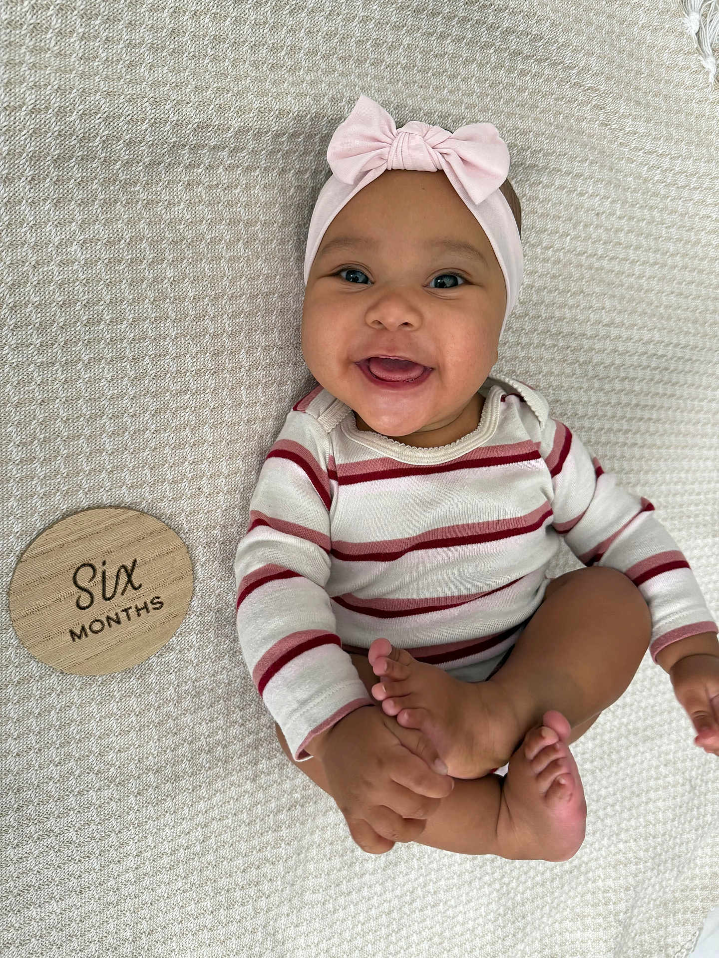 Aria is registered to the contest to win money with this photo: baby, infant, smiling, headband, pink_bow, striped_clothing, sitting, barefoot, hands, feet, wooden_plaque, text, blanket, texture, happy, cute, portrait, child, six_months, indoors