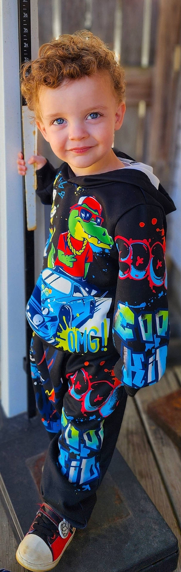 Joseph is registered to the contest to win money with this photo: blue_eyes, boy, cartoon_graphics, casual_wear, child, colorful_clothing, curly_hair, cute, happy, hoodie, metal_pole, outdoor, pants, playful, portrait, smiling, sneakers, standing, urban_style, wooden_floor