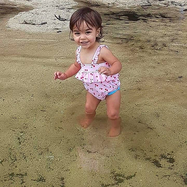 Kallie is registered to the contest to win money with this photo: baby_toddler_clothing, barefoot, beach, body_of_water, eye, foot, fun, hair, happy, head, human_body, human_leg, joy, leg, people_in_nature, people_on_beach, person, sand, smile, thigh
