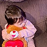 toddler, child, teddy_bear, plush_toy, valentines_day, heart, pink_clothing, couch, furniture, indoor, hugging, smile, cute, soft_toy, person, baby, young_child, sitting, cozy, home