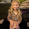 child, girl, young, smiling, portrait, sitting, couch, living_room, leopard_print, pajamas, blonde_hair, hair_bow, green_plush, stuffed_animal, blanket, pillow, toy_box, indoors, cozy, family_home