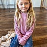 child, girl, portrait, indoors, kneeling, smiling, blonde_hair, long_hair, pink_shirt, jeans, heart_pattern, blanket, wooden_floor, home, living_room, furniture, sitting, face, person, photography