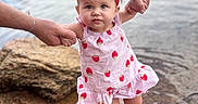 Kennedi is registered to the contest to win money with this photo: adult_hands, baby, barefoot, bow, child, fashion, girl, headband, lakeside, outdoor, parental_support, photography, portrait, rocky_ground, safety, shore, smiling, strawberry_dress, toddler, water