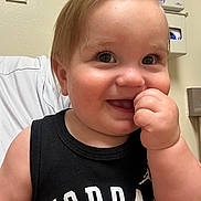 Xander is registered to the contest to win money with this photo: toddler, child, baby, smiling, portrait, close_up, face, blond_hair, blue_eyes, hand, finger_in_mouth, black_shirt, jordan_shirt, sitting, indoors, hospital_room, medical_setting, chair, wall, happy