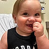 toddler, child, baby, smiling, portrait, close_up, face, blond_hair, blue_eyes, hand, finger_in_mouth, black_shirt, jordan_shirt, sitting, indoors, hospital_room, medical_setting, chair, wall, happy