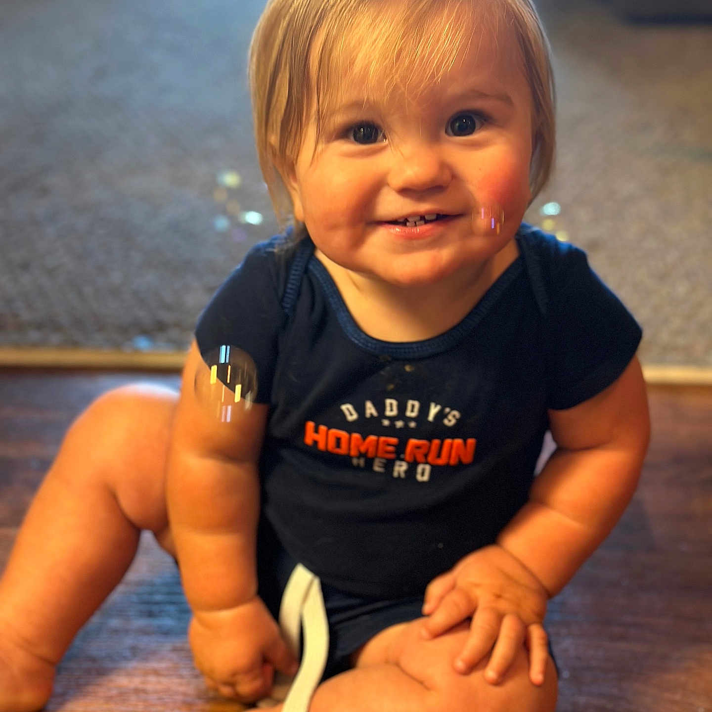 Xander joined the competition — help win amazing prizes! baby, child, toddler, smiling, portrait, indoor, sitting, wooden_floor, baseball_shirt, clothing, blond_hair, blue_eyes, face, hands, legs, happy, home, warm_lighting, photography, infant