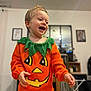 toddler, child, pumpkin_costume, orange_clothing, green_collar, laughing, happy, indoor, table, wooden_table, face, person, smile, short_hair, home, blurred_background, framed_pictures, furniture, cozy, festive