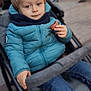 toddler, child, stroller, blue_jacket, scarf, macaron, snack, outdoor, street, people, blurred_background, jeans, casual_clothing, winter_wear, young_child, sitting, holding_food, curious_expression, daylight, urban