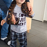 Nino a rejoint le concours — aidez-le/la à gagner de superbes lots ! child, toddler, snake, reptile, indoor, plaid_pants, tshirt, adult_hand, floor_tiles, wooden_furniture, blue_eyes, short_hair, standing, animal, pet, curious, casual_clothing, support, light, portrait