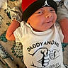 newborn, baby, infant, child, smiling, face, portrait, lying_down, indoors, blanket, clothing, onesie, hat, cap, red, black, white, text, patterned_background, close_up