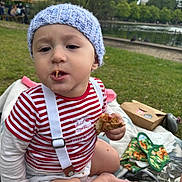 Theo joined the competition — help win amazing prizes! toddler, child, baby, hat, knitted_hat, striped_shirt, suspenders, food, bread, picnic, blanket, grass, park, outdoor, nature, pond, trees, daytime, cute, sitting