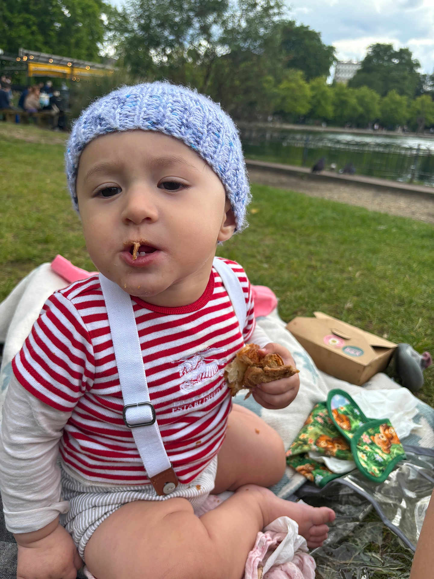 Theo joined the competition — help win amazing prizes! toddler, child, baby, hat, knitted_hat, striped_shirt, suspenders, food, bread, picnic, blanket, grass, park, outdoor, nature, pond, trees, daytime, cute, sitting