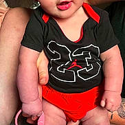 Jesslynn is registered to the contest to win money with this photo: adult, baby, black_shirt, carpet, child, close_up, indoor, infant, portrait, rainbow_rings, red_bow, red_clothing, silver_shoes, smiling, socks, standing, superhero_costume, supported, tattoo, toy