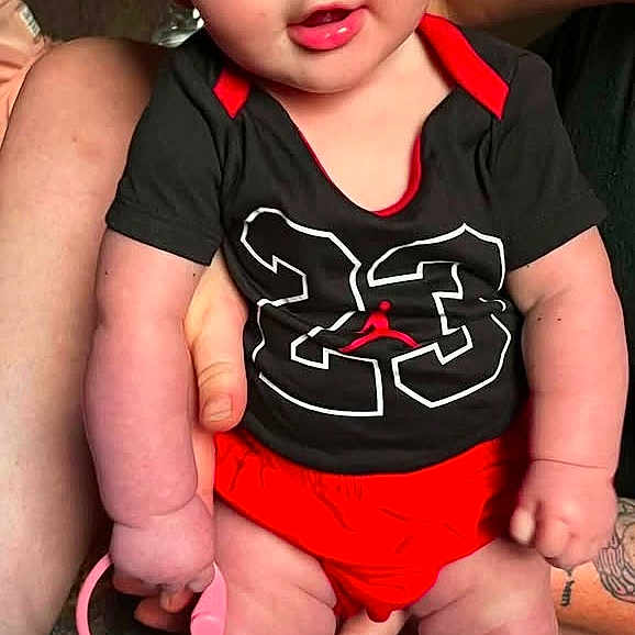 Jesslynn is registered to the contest to win money with this photo: adult, baby, black_shirt, carpet, child, close_up, indoor, infant, portrait, rainbow_rings, red_bow, red_clothing, silver_shoes, smiling, socks, standing, superhero_costume, supported, tattoo, toy