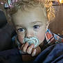toddler, child, pacifier, curly_hair, blue_eyes, indoor, blanket, closeup, portrait, hand, face, young_child, cozy, curly, infant, plaid_shirt, cute, baby, indoor_lighting, soft_light