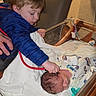 Ronin is registered to the contest to win money with this photo: child, newborn, baby, hospital, bassinet, stethoscope, sibling, blue_jacket, blanket, medical_tube, bedroom_chair, person, infant, cute, family, indoor, healthcare, care, love, tenderness