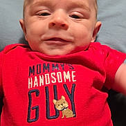 Ronin is registered to the contest to win money with this photo: apparel, baby, blue_blanket, casual_wear, child, closeup, cute, expression, face, head, indoors, infant, lying_down, newborn, person, portrait, red_onesie, smiling, text_on_clothing, young
