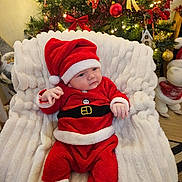 Matéo participe au concours pour gagner de l'argent avec cette photo : baby, christmas, holiday, santa_costume, hat, red_clothing, white_fur_trim, blanket, chair, decorations, tree, ornaments, lights, festive, indoors, child, cute, smiling, seated, celebration