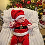 baby, christmas, holiday, santa_costume, hat, red_clothing, white_fur_trim, blanket, chair, decorations, tree, ornaments, lights, festive, indoors, child, cute, smiling, seated, celebration