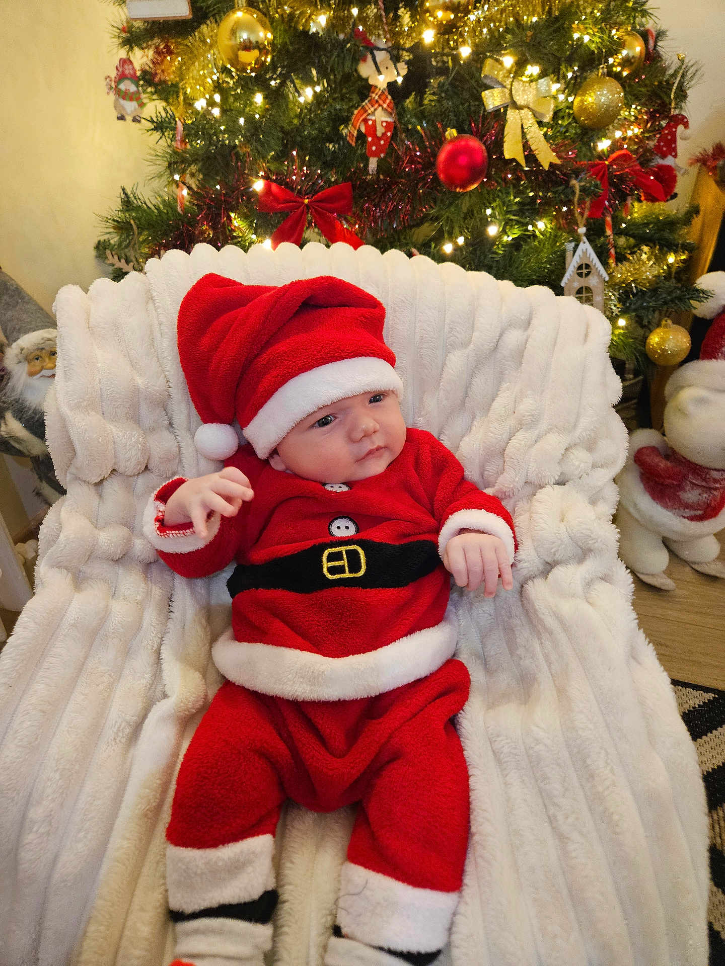 Matéo participe au concours pour gagner de l'argent avec cette photo : baby, christmas, holiday, santa_costume, hat, red_clothing, white_fur_trim, blanket, chair, decorations, tree, ornaments, lights, festive, indoors, child, cute, smiling, seated, celebration