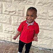 Andre is registered to the contest to win money with this photo: toddler, child, smile, red_shirt, white_sneakers, standing, outdoor, stone_wall, happy, portrait, cute, young, person, casual_clothing, black_pants, short_hair, daylight, full_body, joyful, fashion