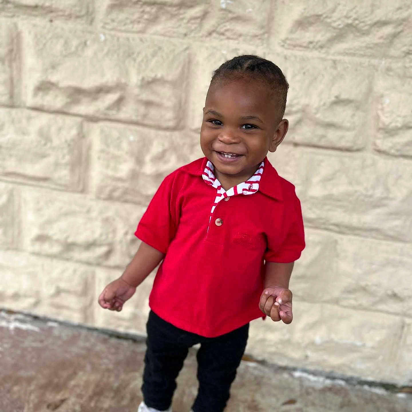 Andre is registered to the contest to win money with this photo: black_pants, casual_clothing, child, cute, daylight, fashion, full_body, happy, joyful, outdoor, person, portrait, red_shirt, short_hair, smile, standing, stone_wall, toddler, white_sneakers, young