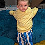 child, toddler, yellow_sweater, striped_pants, blue_and_white_stripes, sofa, couch, blanket, pillow, indoor, portrait, standing, smiling, eyes, hair, cute, home_interior, fabric_texture, childrens_clothing, playful