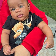 Namir is registered to the contest to win money with this photo: baby, child, person, holding, black_clothing, teddy_bear, necklace, red_hoodie, hand, outdoor, grass, sidewalk, parked_cars, curious_expression, casual, portrait, infant, cute, relaxed, daylight