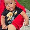 baby, child, person, holding, black_clothing, teddy_bear, necklace, red_hoodie, hand, outdoor, grass, sidewalk, parked_cars, curious_expression, casual, portrait, infant, cute, relaxed, daylight