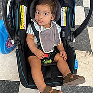 Chris is registered to the contest to win money with this photo: baby, bib, car_seat, casual_clothing, checkered_floor, child, curious, floor, footwear, indoor, person, portrait, safety, sandals, seat, seat_belt, striped_shirt, toddler, warning_label, young_child