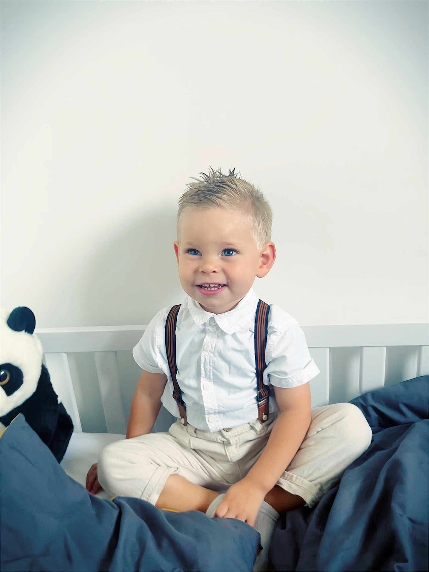 Charly a rejoint le concours — aidez-le/la à gagner de superbes lots ! child, boy, smiling, bed, panda_plush, white_shirt, suspenders, blonde_hair, blue_eyes, pants, cross_legged, indoors, cute, happy, portrait, soft_lighting, plush_toy, furniture, casual_clothing, young