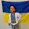 Bogdan a rejoint le concours — aidez-le/la à gagner de superbes lots ! child, boy, standing, traditional_clothing, embroidered_shirt, flag, blue, yellow, wall, expression, playful, indoors, casual, pants, curly_hair, mischievous, person, portrait, cultural, fun