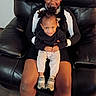 Melvin is registered to the contest to win money with this photo: child, toddler, sibling, smiling, portrait, indoor, leather_chair, seated, holding, family, black_clothing, headband, braids, baby, white_pants, sneakers, furniture, home, happy, photograph