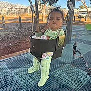 Jayden is registered to the contest to win money with this photo: baby, black_swing, blanket, child, daylight, dinosaur_print, green_clothing, infant, metal_chains, nature, outdoor, pajamas, parked_cars, person, play_surface, playground, seat, serious_expression, swing, trees