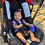 baby, infant, smile, car_seat, stroller, purple_toy, harness, bow_in_hair, bare_feet, purple_shirt, sunlight, outdoor, dirt_ground, safety_label, canopy, black_cushion, happy, portrait, seatbelt, child