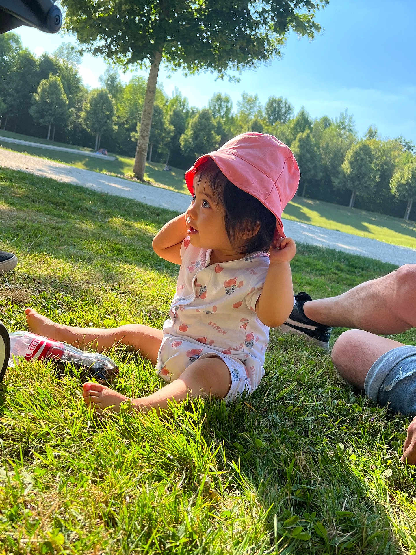 June a rejoint le concours — aidez-le/la à gagner de superbes lots ! toddler, child, pink_hat, outdoor, grass, sunlight, park, tree, blue_sky, coca_cola_bottle, casual_clothing, shorts, sneakers, daylight, nature, sitting, curious, cute, person, summer