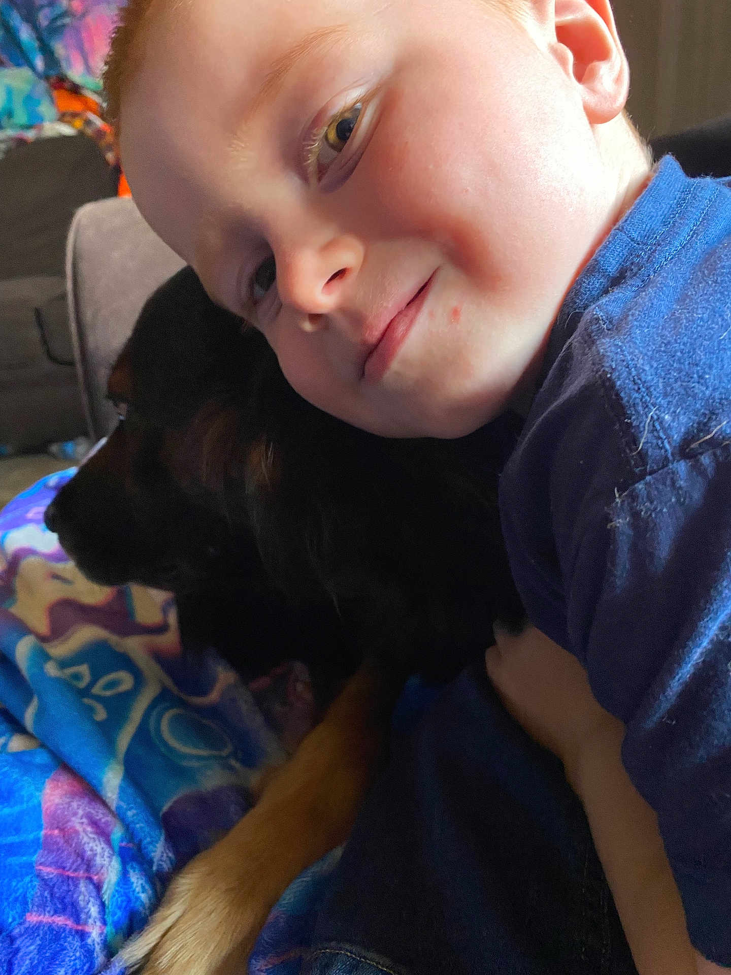 James joined the competition — help win amazing prizes! child, dog, cuddle, smile, face, blanket, blue_shirt, pet, indoor, cozy, happy, animal, friendship, comfort, closeup, portrait, young_child, furniture, relaxation, domestic