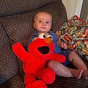 Tommy joined the competition — help win amazing prizes! baby, child, plush_toy, elmo, couch, pillow, indoor, smile, cute, seated, clothing, footwear_pattern, red, blue, toy, person, face, comfort, home, expression