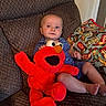 baby, child, plush_toy, elmo, couch, pillow, indoor, smile, cute, seated, clothing, footwear_pattern, red, blue, toy, person, face, comfort, home, expression