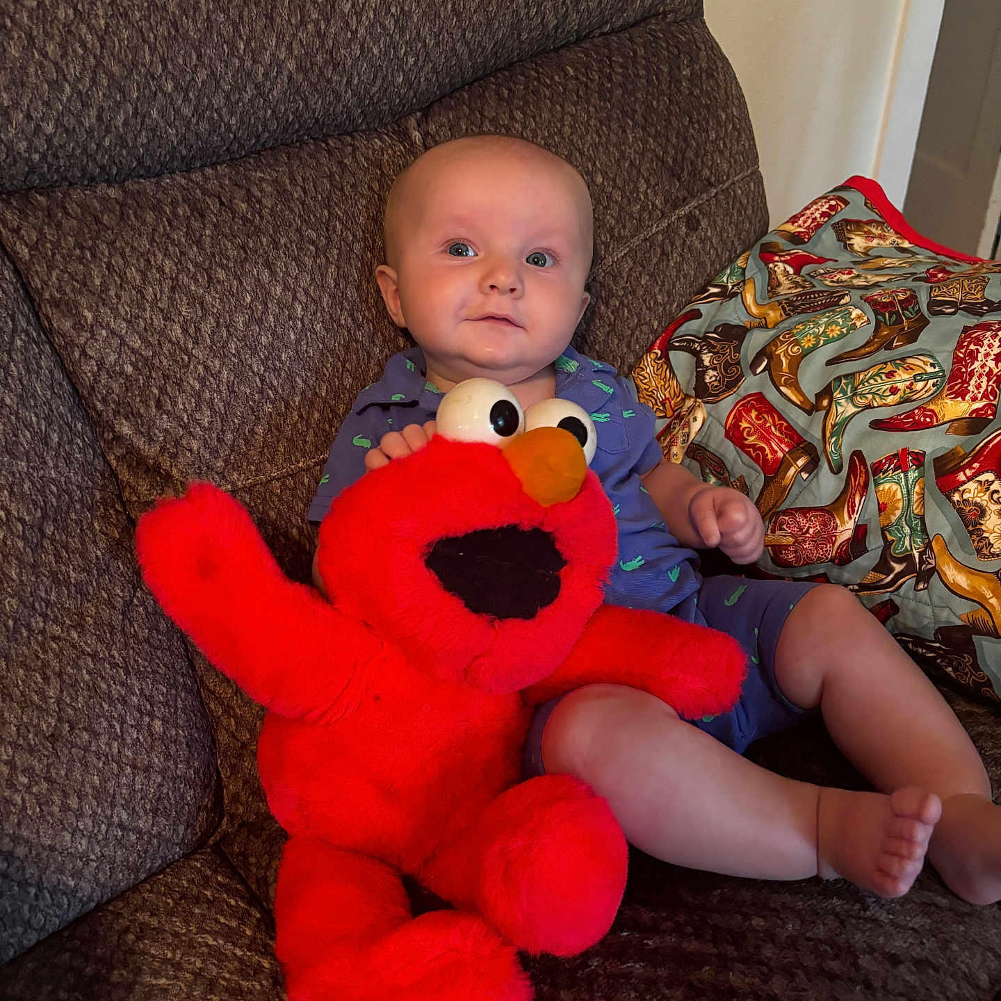 Tommy joined the competition — help win amazing prizes! baby, child, plush_toy, elmo, couch, pillow, indoor, smile, cute, seated, clothing, footwear_pattern, red, blue, toy, person, face, comfort, home, expression