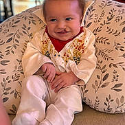 Sean is registered to the contest to win money with this photo: baby, child, clothing, cozy, cute, embroidery, face, feet, hands, indoors, infant, patterned, person, pillow, portrait, red_bib, sitting, smiling, soft_lighting, white_outfit
