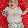 baby, infant, child, lying_down, red_quilt, floral_pattern, white_shirt, giraffe_print, colorful_shorts, cute, blue_eyes, hand, floor, playmat, portrait, indoors, smiling, young_child, clothing, baby_fashion